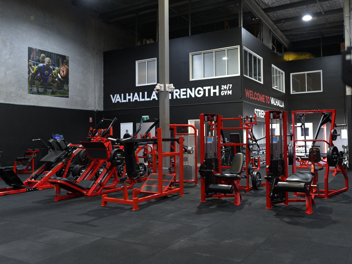 A gym interior with multiple red and black strength training machines arranged in a large black-and-gray facility. Visible wall signage reads 'VALHALLA STRENGTH 24/7 GYM' and 'WELCOME TO VALHALLA'.